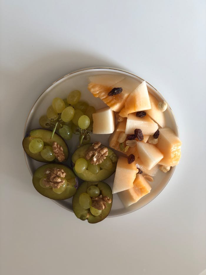 A vibrant, healthy fruit and nut platter featuring grapes, melon, plums, and walnuts on a white background.